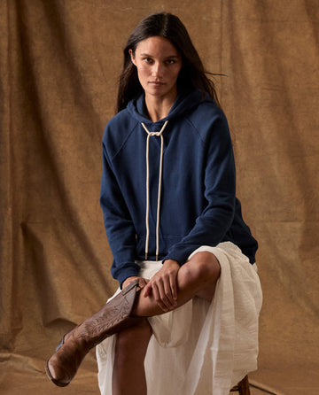 A person wearing a True Navy The Fleece Pullover Hoodie with thick white drawstrings, a white skirt, and brown cowboy boots.