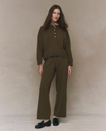 A woman stands against a neutral backdrop in THE GREAT. F25 HIGH TWIST The Henley Sweater—Army, paired with matching wide-leg pants, a patterned scarf, and black shoes for a relaxed menswear-inspired look.