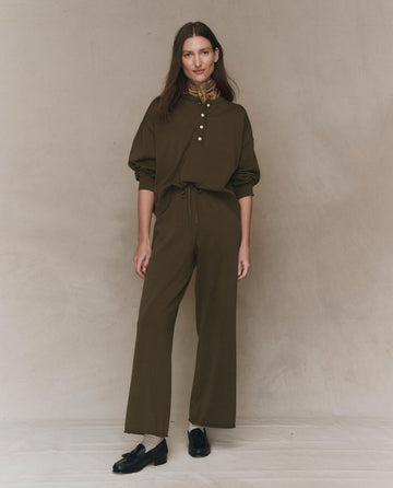 A woman stands against a neutral backdrop wearing THE GREAT. F25 HIGH TWIST The Rec Sweater Pant in Army with an elastic waistband, styled with a loose olive-green shirt and black shoes, looking at the camera in a relaxed pose.