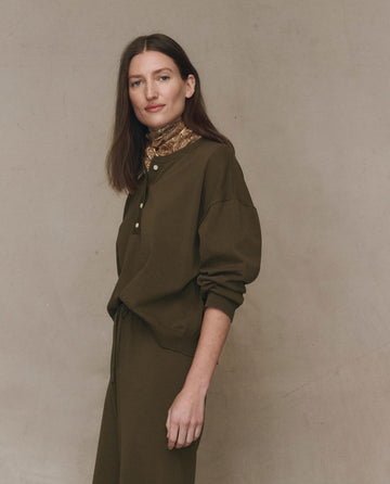 A woman with long brown hair stands against a plain background, wearing THE GREAT. F25 HIGH TWIST The Henley Sweater in Army and a lightweight knit scarf around her neck.