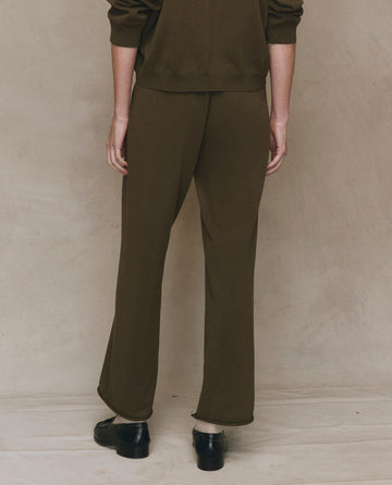 A person stands facing away from the camera, wearing THE GREAT. F25 HIGH TWIST The Rec Sweater Pant in Army with an elastic waistband, paired with black shoes against a neutral background.