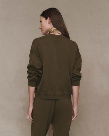 A woman with long brown hair wears THE GREAT. F25 HIGH TWIST The Henley Sweater -- Army, standing with her back to the camera against a beige wall.