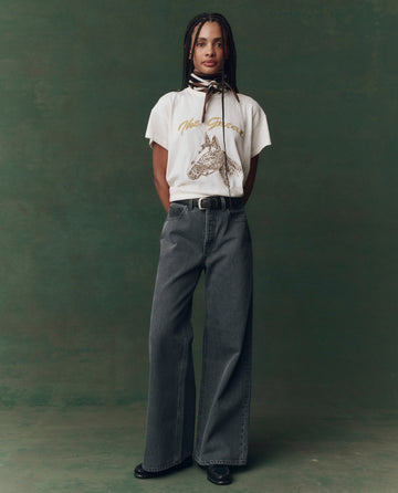 A person stands against a green backdrop wearing THE GREAT. F25 D2's The Boxy Crew. Embroidered in washed white with spice horse embroidery, paired with wide-leg gray jeans, black shoes, and a striped scarf around their neck.