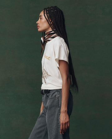 A person with long braided hair stands in profile against a dark green background, wearing THE GREAT.'s The Boxy Crew. Embroidered tee in washed white with spice horse embroidery.