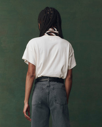 A person faces away, wearing THE GREAT. F25 D2 The Boxy Crew Embroidered in Washed White with Spice Horse Embroidery, paired with high-waisted gray jeans and a black-and-white scarf, against a plain green background.