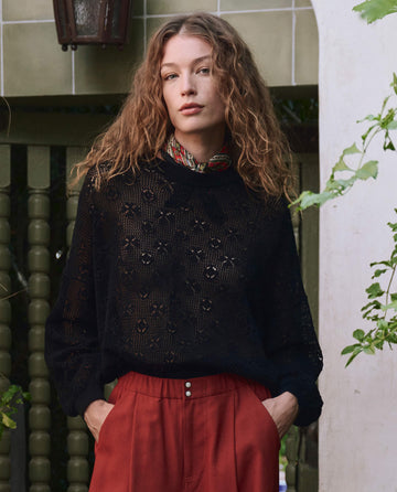 A person with wavy hair wears THE GREAT. PF25 The Crochet Pullover in black over a lace top, styled with red trousers and a patterned neck scarf, standing outdoors by a green fence and tiled wall.