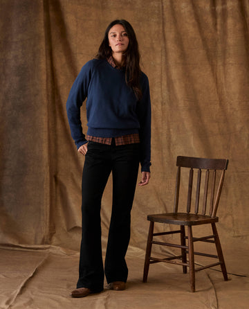 A person wearing a navy blue THE FLEECE ROOKIE SWEATSHIRT over a plaid collared shirt and black flared pants, standing next to a wooden chair.