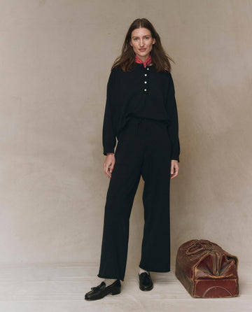 A woman wearing THE GREAT. F25 HIGH TWIST The Rec Sweater Pant in black and a coordinating black long-sleeve top with a red patterned collar stands next to a brown vintage travel bag against a neutral background.