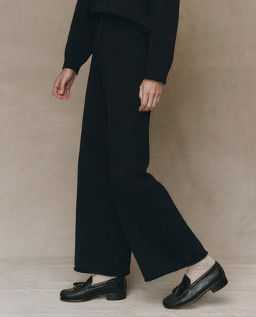 A person in THE GREAT. F25 HIGH TWIST The Rec Sweater Pant in black stands against a plain beige background, wearing a black long-sleeve top, light socks, and black loafers.