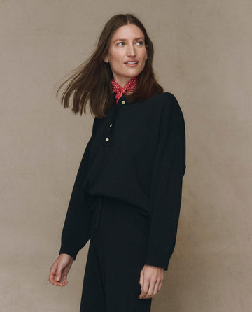 A woman with straight brown hair wears THE GREAT. F25 HIGH TWIST The Henley Sweater in black and a red patterned scarf, standing against a plain beige background.