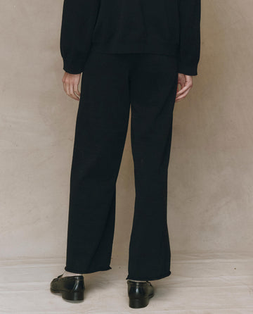 Person standing against a beige wall, shown from the waist down, wearing THE GREAT. F25 HIGH TWIST The Rec Sweater Pant in black paired with black shoes and a black long-sleeve top.