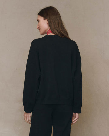 A person with long brown hair, wearing THE GREAT.’s The Henley Sweater in black (F25 HIGH TWIST) and black pants, stands facing away from the camera against a neutral background.