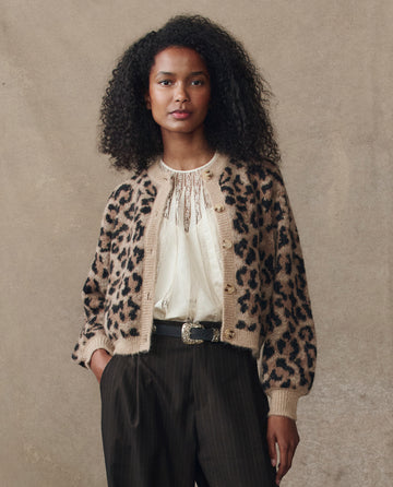 A woman with curly hair wears THE GREAT. H25 COLLECTION's The Windsor Cardigan in Leopard over a light blouse and dark pinstripe trousers, standing against a plain beige background.