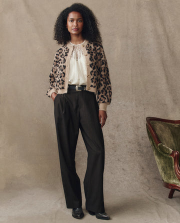 A woman stands indoors wearing THE GREAT. H25 COLLECTION’s The Windsor Cardigan in leopard, a cream blouse, dark high-waisted trousers, and black shoes, one hand in her pocket. Beside her is an empty green velvet chair.