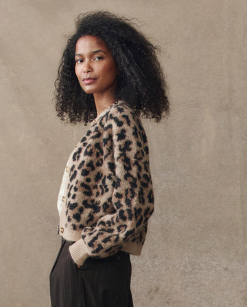 A woman with curly hair wears THE GREAT. H25 COLLECTION's The Windsor Cardigan in Leopard with dark pants, standing against a beige background and facing slightly sideways.