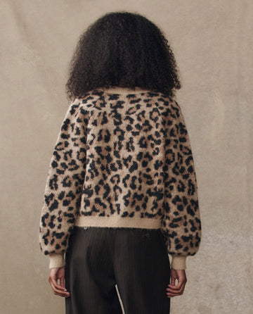Someone with curly hair faces away, wearing THE GREAT. H25 COLLECTION’s The Windsor Cardigan in Leopard print and dark pants, set against a plain beige background.