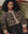 A person with curly hair sits on a green sofa, wearing THE GREAT. H25 COLLECTION’s The Windsor Cardigan in leopard print and dark pinstriped pants.