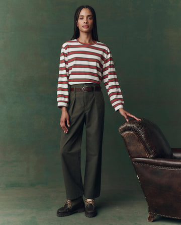 A person stands indoors beside a brown leather chair, wearing THE GREAT. F25 D2’s The Campus Crew. Novelty in Cream Bridle Stripe, dark green trousers, and brown shoes against a muted green background.