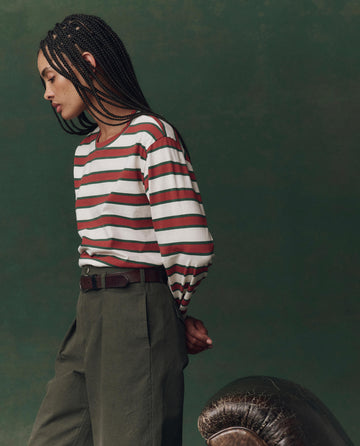 A person with braided hair in profile wears THE GREAT. F25 D2’s The Campus Crew. Novelty -- Cream Bridle Stripe with green pants, standing by a brown leather chair against a green backdrop for a relaxed, campus-inspired style.