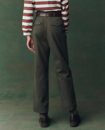 Person stands with their back to the camera wearing THE GREAT. F25 D2 The Parker Trouser in Dusty Moss, a striped long-sleeve shirt, brown belt, and dark brown boots against a green background.