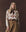A woman with long blonde hair wears THE GREAT. The Slouch Sweatshirt in the Riding Horses Print, styled with a brown scarf and belted tan skirt, standing against a brown background.