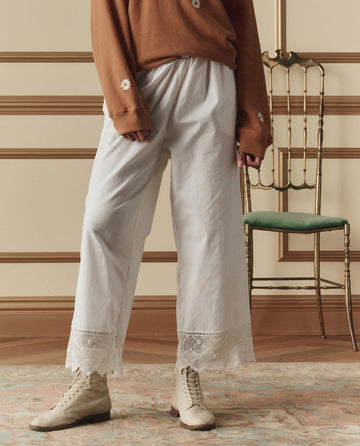A person wearing a brown long-sleeve shirt, THE GREAT. SP26 D2 The Rosette Crochet Pant in cream, and beige lace-up boots stands indoors near a gold chair with a green cushion.