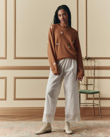 Indoors, a woman wears THE GREAT. SP26 D2’s The Rosette Crochet Pant in cream, paired with a brown floral sweater and beige boots; a green chair sits in the background.
