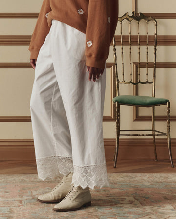 A person stands indoors wearing THE GREAT. SP26 D2 The Rosette Crochet Pant in cream, paired with a brown embroidered flower sweater and cream lace-up shoes. An ornate chair with a green seat appears in the background.