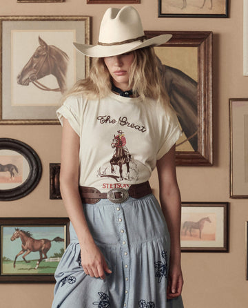 A woman in a cowboy hat and western outfit wears THE GREAT. STETSON SP26 "The Tooled Waist Belt" in brown with silver, standing before a wall adorned with framed horse artwork.