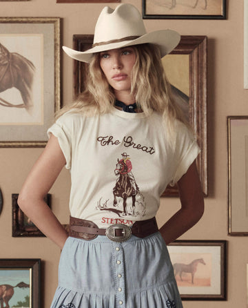 A woman wearing THE GREAT. STETSON SP26’s The Tooled Waist Belt in Brown with Silver stands before a wall adorned with framed horse pictures, pairing it with a cowboy hat, graphic tee, and skirt.