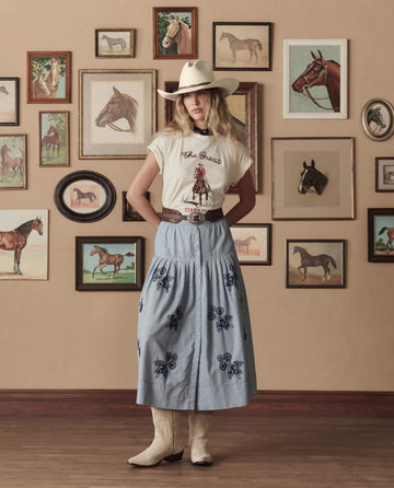 A woman in THE GREAT. STETSON SP26 The Boxy Crew With Stetson Graphic in Washed White, a cowboy hat, embroidered skirt, and boots stands before a wall decorated with framed horse pictures.