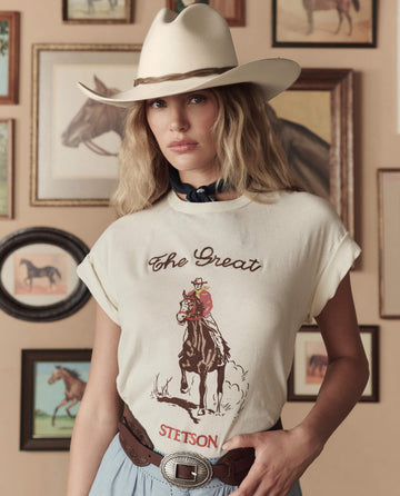 A woman in a cowboy hat, THE GREAT. STETSON SP26 t-shirt, and The Tooled Waist Belt—Brown With Silver stands before a wall decorated with framed horse paintings.
