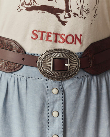 Close-up of a person wearing a beige STETSON SP26 shirt and The Tooled Waist Belt in brown with silver by THE GREAT., paired with a blue buttoned skirt.