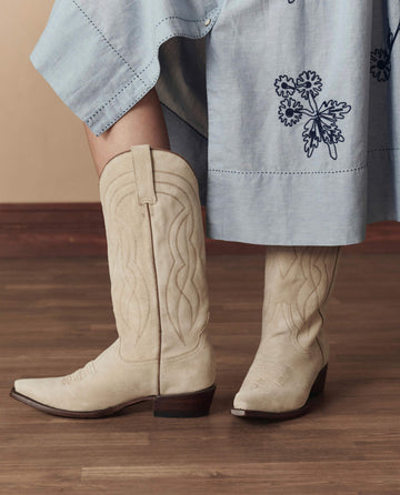 A person wearing THE GREAT. STETSON SP26 "The Cowboy Boot (Suede)" in Pearl stands on a wooden floor, paired with a light blue skirt featuring dark blue floral embroidery.