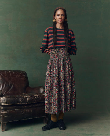 A woman stands beside a worn leather armchair, wearing The Campus Crew. Novelty -- Navy Bridle Stripe top from THE GREAT. F25 D2, a patterned skirt, headband, and black shoes, set against a green backdrop.