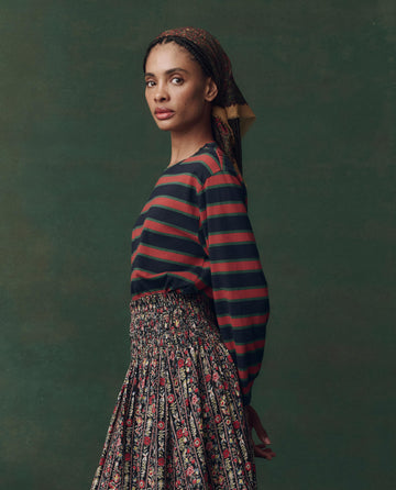 A woman in profile poses against a green backdrop wearing THE GREAT. F25 D2’s The Campus Crew. Novelty — Navy Bridle Stripe, styled with a patterned skirt and headscarf for a relaxed made-in-L.A. vibe.