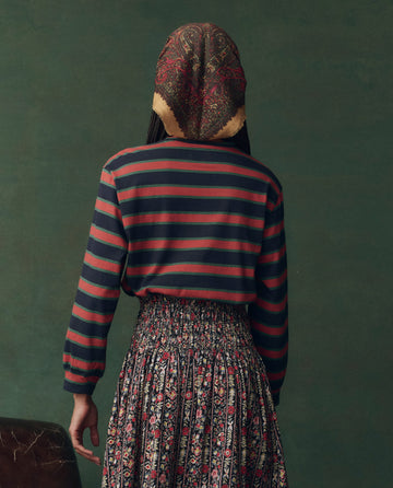 A person faces away in THE GREAT. F25 D2’s The Campus Crew. Novelty—Navy Bridle Stripe shirt, paired with a floral skirt and patterned scarf, set against a plain green background for an effortlessly stylish L.A.-made look.