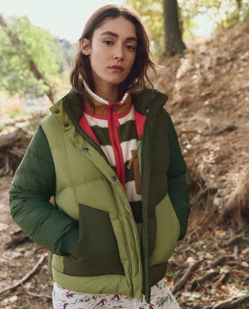 A woman stands outdoors in THE GREAT. F25 TGO The Polar Puffer -- Army Colorblock, featuring PrimaLoft Black Thermo Plume insulation, layered over a fleece and patterned pants, with sunlight and trees in the background.