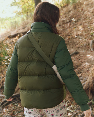 A person wearing THE GREAT. F25 TGO The Polar Puffer -- Army Colorblock jacket with PrimaLoft Black Thermo Plume insulation and patterned pants walks outdoors on a dirt path surrounded by dry grass and trees.