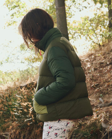 A person in THE GREAT. F25 TGO The Polar Puffer—Army Colorblock with PrimaLoft Black Thermo Plume insulation and patterned pants stands outdoors in a wooded area, hands in pockets, facing sideways.