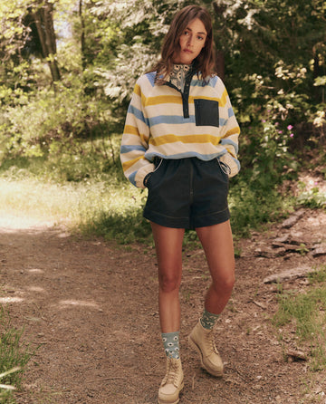 A person stands on a forest path wearing The Excursion Pullover -- Meadow Stripe by THE GREAT. SP25 TGO, dark shorts, patterned socks, and beige boots, surrounded by trees and vibrant foliage.