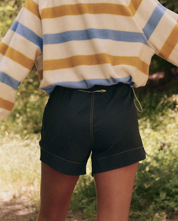 A person in a striped sweater and The Rover Short (Navy) by THE GREAT. SP25 TGO stands outdoors with arms extended, surrounded by lush greenery.