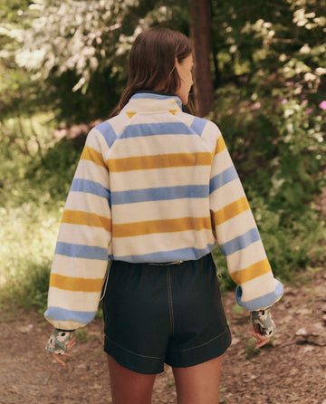 A person with long hair, wearing a Meadow Stripe Excursion Pullover from THE GREAT. SP25 TGO and dark shorts, walks along a wooded path, facing away.