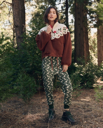A woman stands in a forest wearing The Plush Wildflower Pullover. -- Chestnut by THE GREAT. F26 TGO, paired with green patterned pants and boots among tall trees.