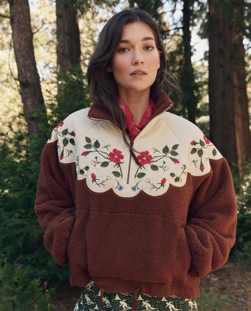 Woman outdoors by trees, wearing THE GREAT. F26 TGO The Plush Wildflower Pullover. -- Chestnut with a cream floral embroidered yoke and a red neck scarf.