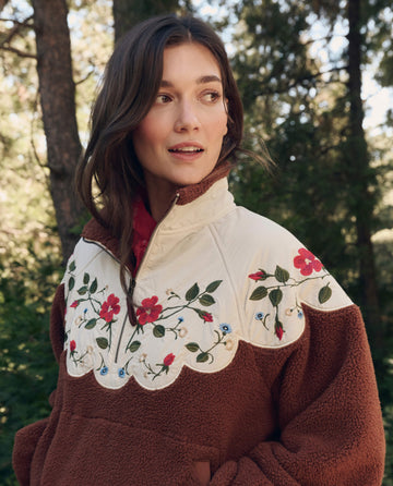 Woman outdoors in The Plush Wildflower Pullover. -- Chestnut by THE GREAT. F26 TGO, featuring a cream scalloped yoke and colorful floral embroidery; trees in background.