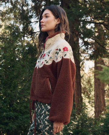 A woman stands among trees, wearing THE GREAT. F26 TGO The Plush Wildflower Pullover. -- Chestnut with floral embroidery and patterned green pants, facing left with a neutral expression.