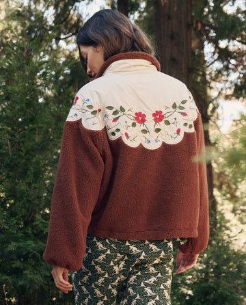 A person outdoors wears THE GREAT. F26 TGO The Plush Wildflower Pullover. -- Chestnut with floral embroidery on the back and patterned green pants, trees in the background.