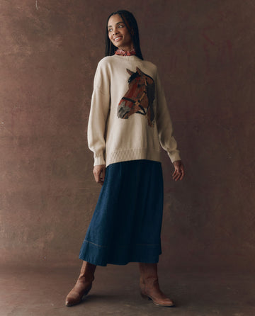 A person smiles, wearing THE GREAT. F25 D1 The Stable Pullover in natural, a cream oversized sweater with a horse-head intarsia, layered over a floral blouse, styled with a long denim skirt and brown boots against a textured brown backdrop.
