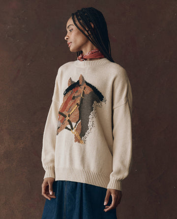 A person wears THE GREAT. F25 D1 The Stable Pullover in Natural, featuring an oversized crew fit and bold horse-head intarsia graphic, standing against a dark brown background and looking to the side.